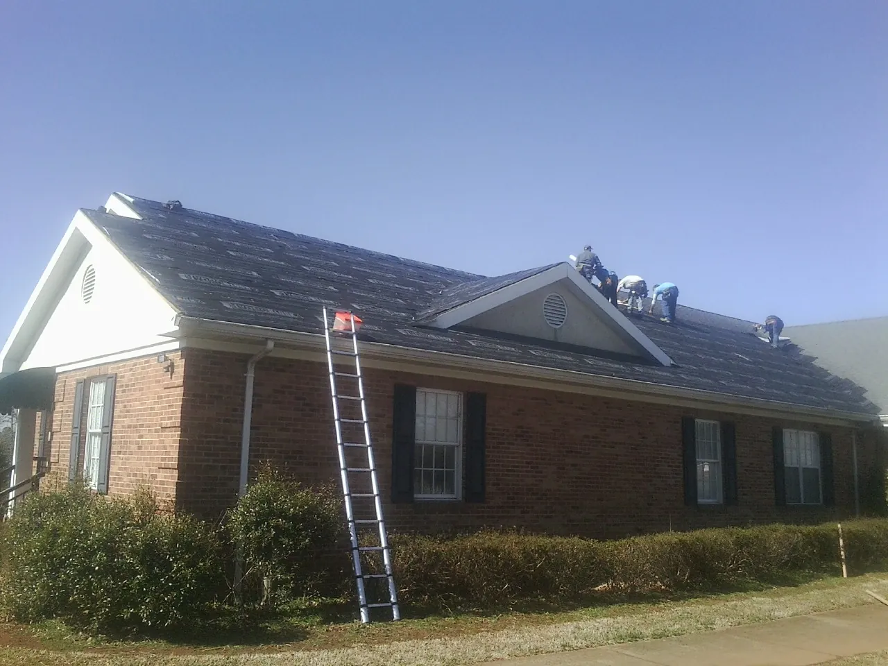 Crew tearing shingles off a brick ranch roof with a ladder set — Emergency Roof Repair work by our crew in Brick