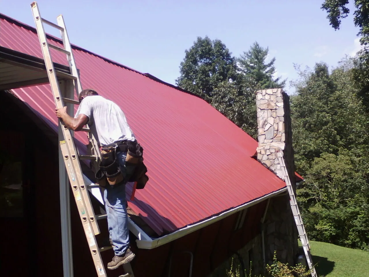 Roofer on a ladder beside a red metal roof and stone chimney — Soffit & Fascia Repair by Roofing Brothers in Brick